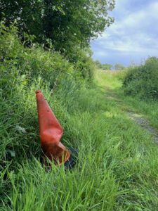 Photo of a crumpled orange plastic traffic cone in some long grass next to a grassy path in a field. There is a hedge behind it. Blue sky in the top right hand corner.