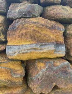 A close up photo of a dry stone wall. The stones are in various shades of copper, brown, and black. The central stone has a darker band running along the bottom which looks like the sea, and above it a copper sunset sky with a dark black cloud on the horizon to the right.