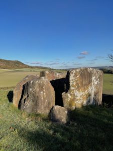 Photo of the megalithic burial site, Coldrum Long Barrow. Four large stones are in the shape of a rectangle, and there is a smaller one on a corner. In the distance is a field and then the Kent Downs. The sky is blue and mostly clear.