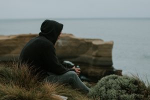 Photo of a man in a hoodie and jeans sat on a grassy cliff top looking at the sea.
