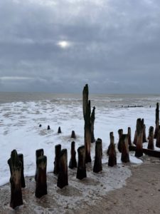 Seaside photo of a grey, cloudy sky with a weak, winter sun behind the clouds. The sea is frothy and browny-grey and lapping at some old, sea-worn, brown wooden posts sticking up from the sand in rows.