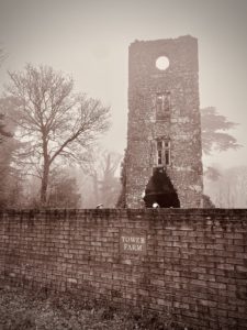 Sepia photo of an old brick tower behind a boundary wall with a Tower Farm sign. The tower has a circular window at the top, two rectangular windows below, and at the bottom a broken wall in the shape of an oval appears above the top of the boundary wall. In the background and to the left of the tower are wintery trees.