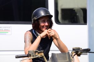 Photo of me in a black tank top with a black half helmet on. I am a white female, with red-brown hair. I am smiling and my elbows are resting on my knees, my hands together and close to my face. You can just see the handlebars of a quad bike.