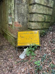Photo of a yellow road sign with a few jumbled numbers and letters. It is leant up against the side of a brick bridge and there are lots of dead leaves on the ground.