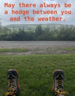 Photo of my hiking boot clad feet, some rolling hills, and text which says: may there always be a hedge between you and the weather.